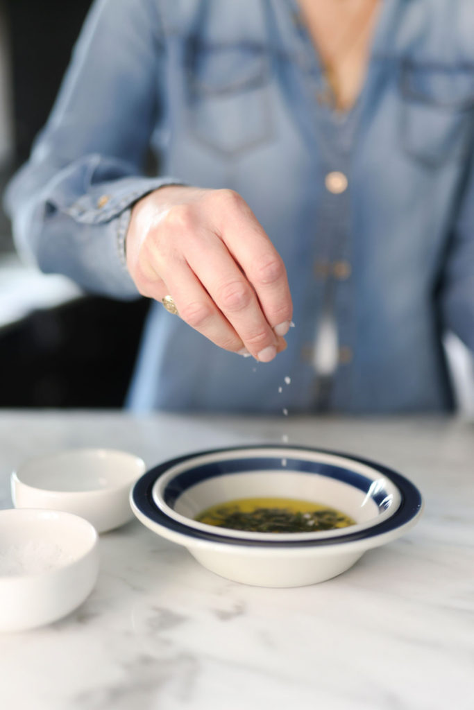 person adding salt in a bowl with olive oil and chopped herbs